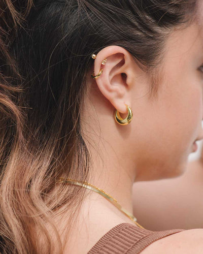A woman wearing the Hailey Hoops, chunky yet elegant gold hoop earrings, plus the Phoebe Studs, Betty Huggies, and two gold necklaces