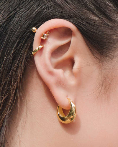 A close up shot of the Hailey Hoops, chunky yet elegant gold hoop earrings, plus the Phoebe Studs and Betty Huggies worn on a woman's right ear