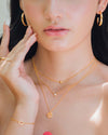 A woman wearing the Sofia Necklace layered with another gold necklace, plus gold hoops, gold crystal studs, gold rings, and a gold bracelet
