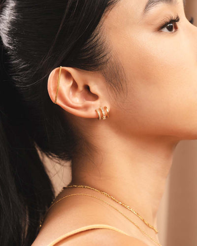 A woman wearing a triple-stacked gold illusion earring, a gold industrial cuff, and 2 gold necklaces
